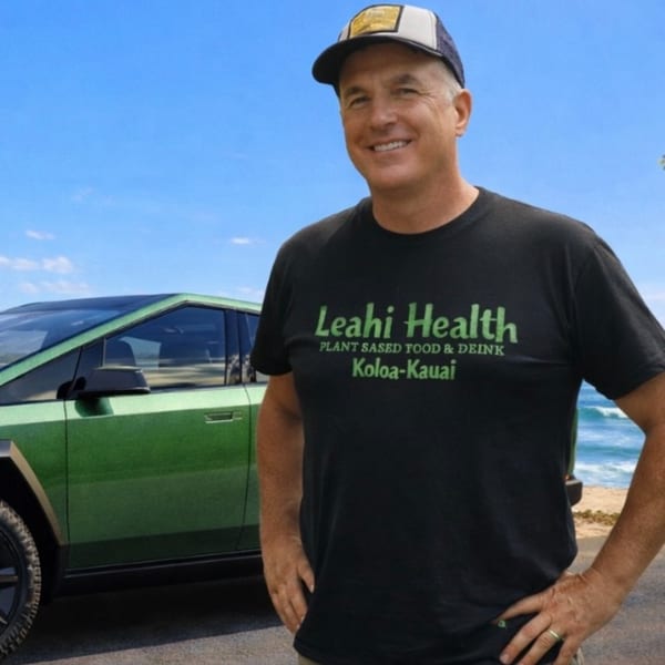 Man in a Leahi Health t-shirt and cap smiling, standing next to a green car by the ocean.