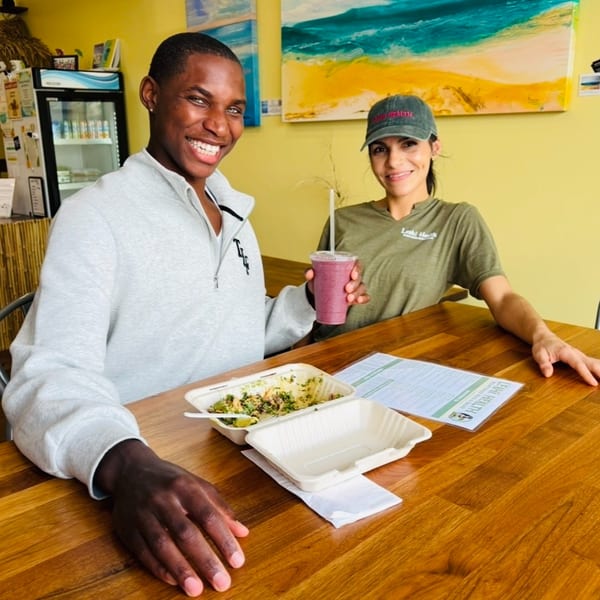 Two smiling people, one holding a smoothie, with a healthy meal on a wooden table at Leahi Health.