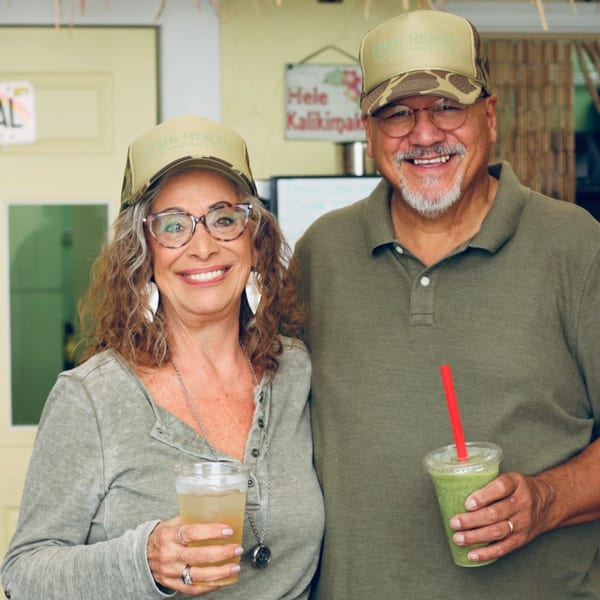 Two smiling Leahi Health employees in branded hats hold healthy drinks, a green smoothie and a light beverage.